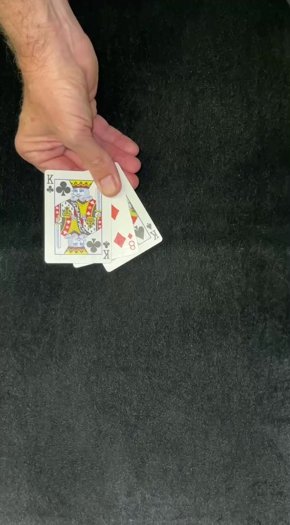 Person's hand holds three playing cards, a King of Clubs, a King of Spades, and an Eight of Diamonds, against a dark surface.