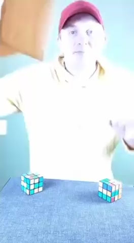 Person in a red cap manipulating two colorful Rubik's cubes on a light-blue surface.