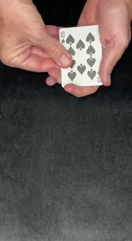 Hands holding a playing card, showing the ten of spades, on a dark surface.