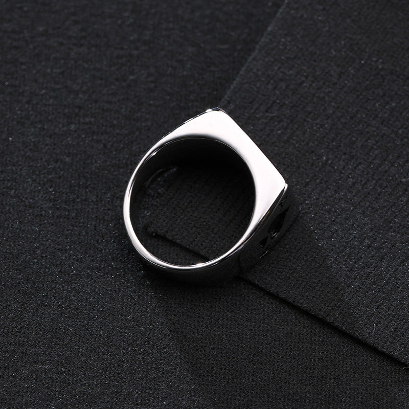 Silver ring with a square top and carved design, resting on black fabric.