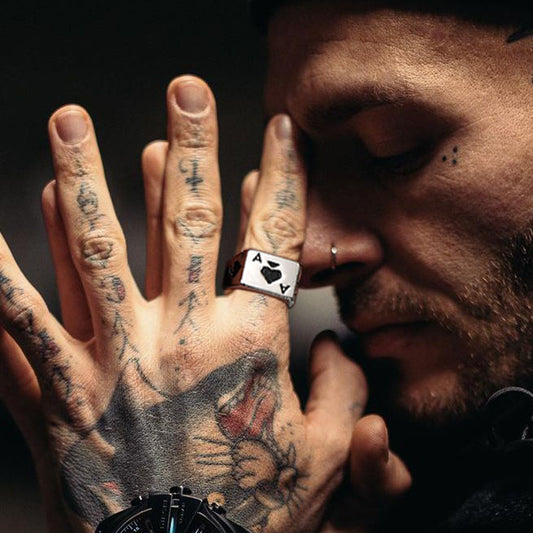 A person with tattoos on their hands and face, wearing an ace of spades ring, rests their hand against their face with eyes closed, creating an introspective mood.